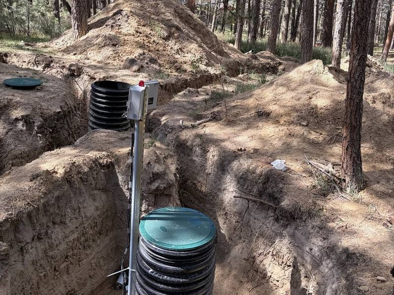 Black corrugated septic riser with a green lid in an open excavation among pine trees